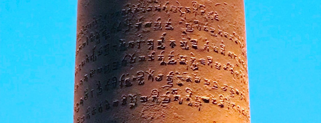 Iron pillar with a Sanskrit inscription written in Brahmi script, set against a clear blue sky.  