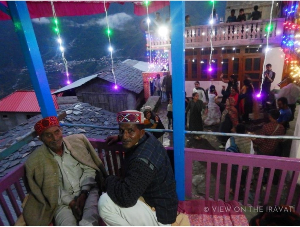 Two Gaddi men sitting on a balcony as people dance during wedding.
